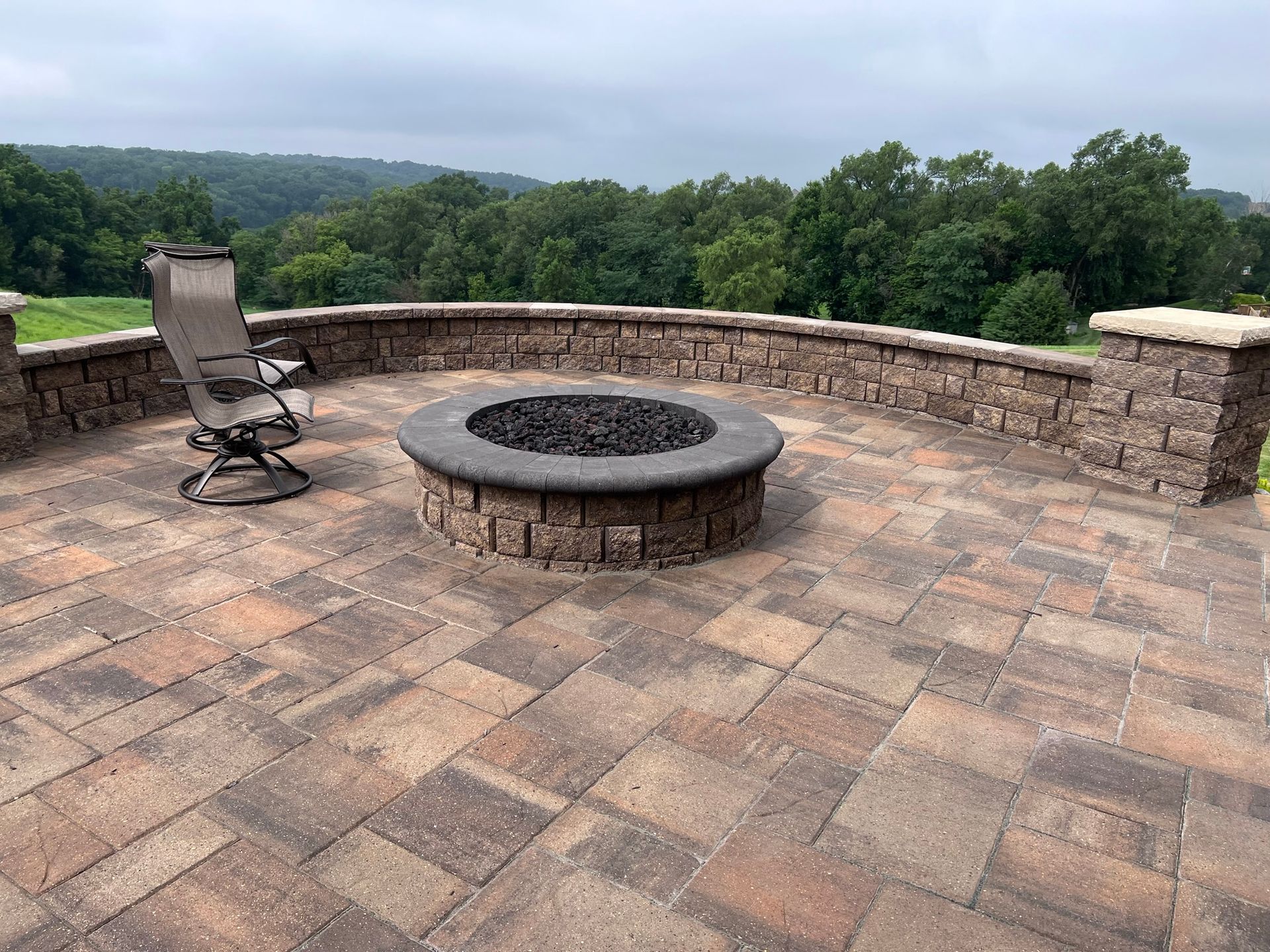 Stone patio with fire pit, seating, and scenic view of trees under cloudy sky.