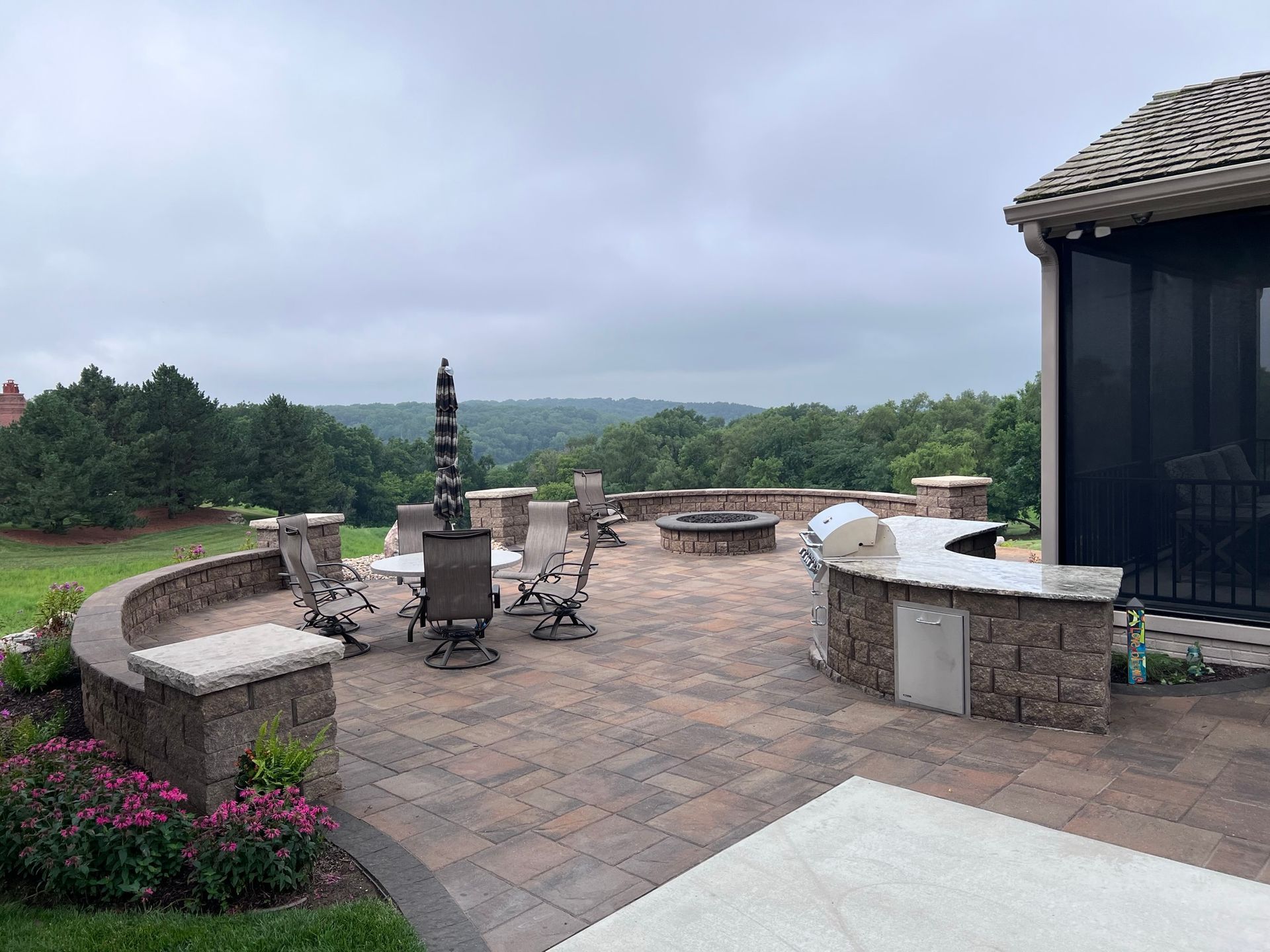Outdoor patio with seating, fire pit, grill, and a scenic view of trees under a cloudy sky.