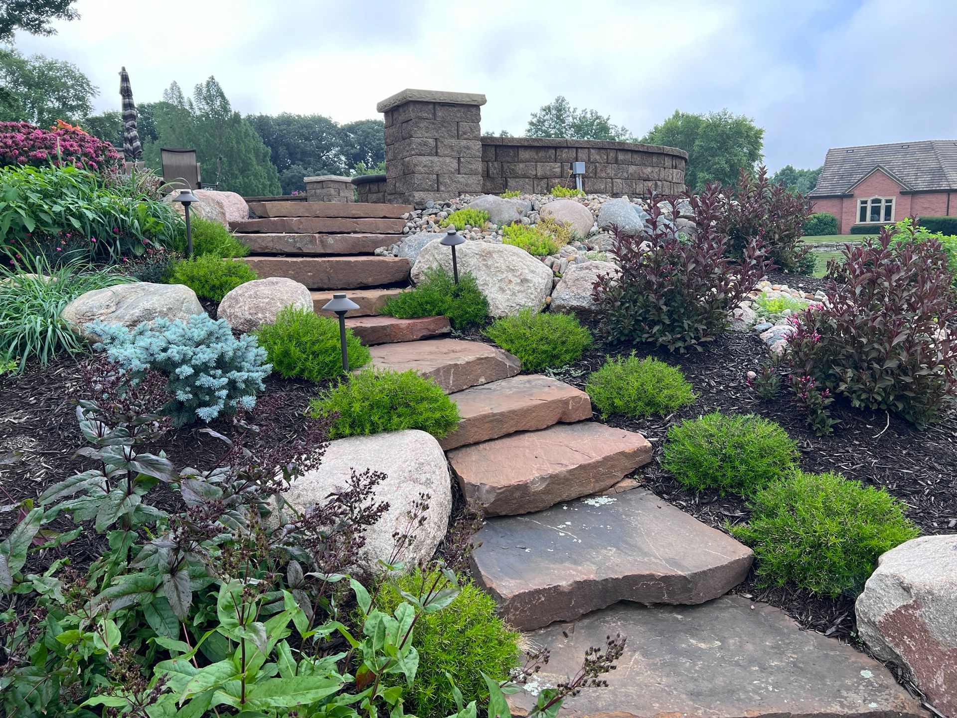 Stone steps ascend a landscaped garden with boulders, shrubs, and a stone structure.
