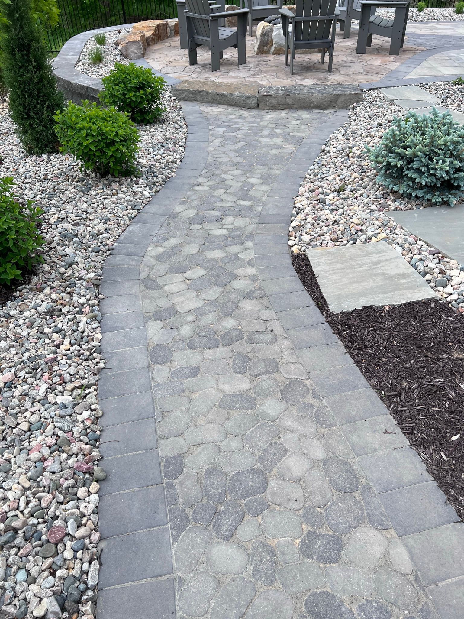 Stone pathway curving through a landscaped yard, leading to a seating area with chairs and a fire pit.