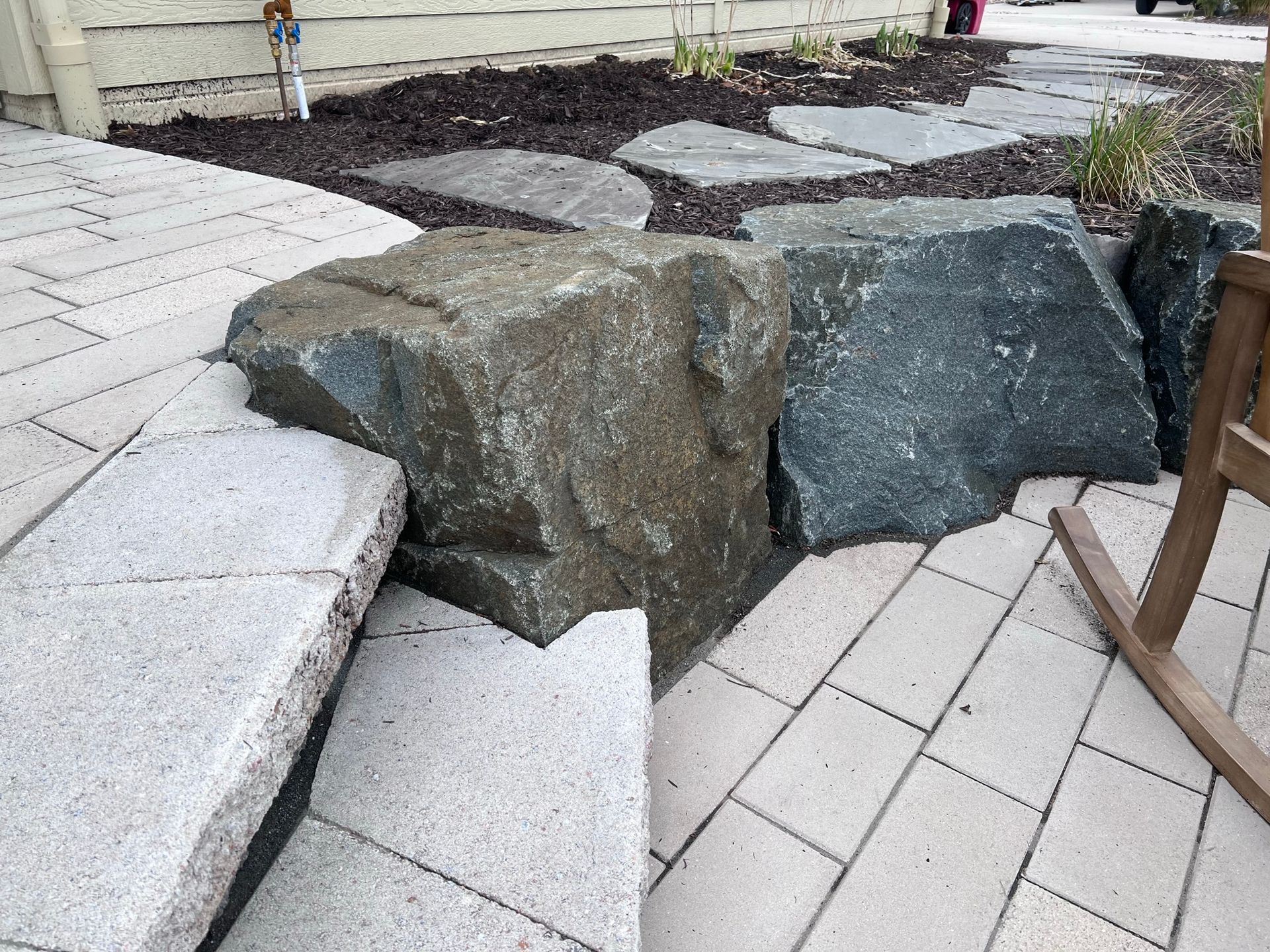 Large gray rock sits beside paved path and flagstone walkway in a garden setting.