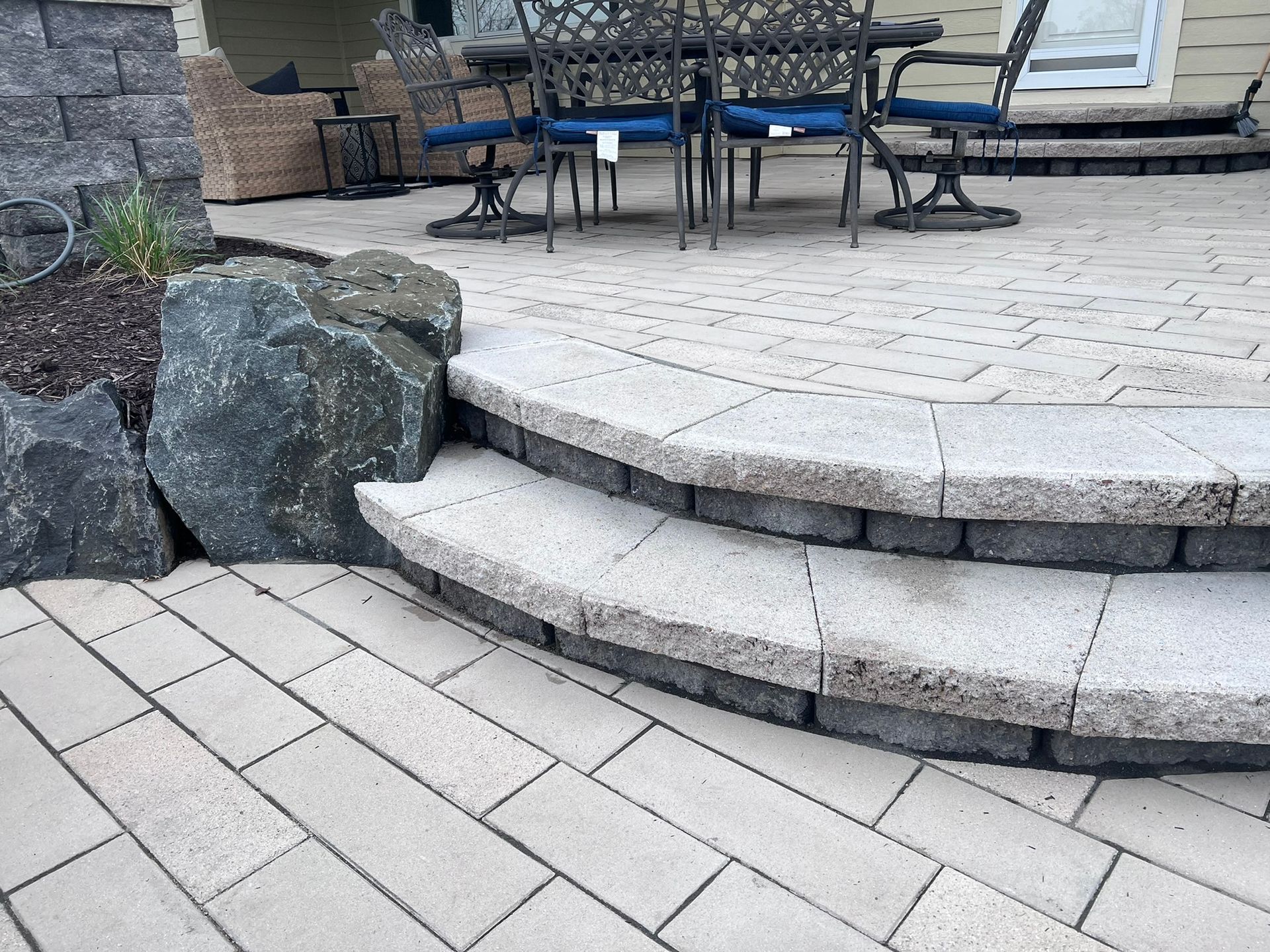 Stone patio with two steps, black edging, large rock feature. Outdoor setting with table and chairs.
