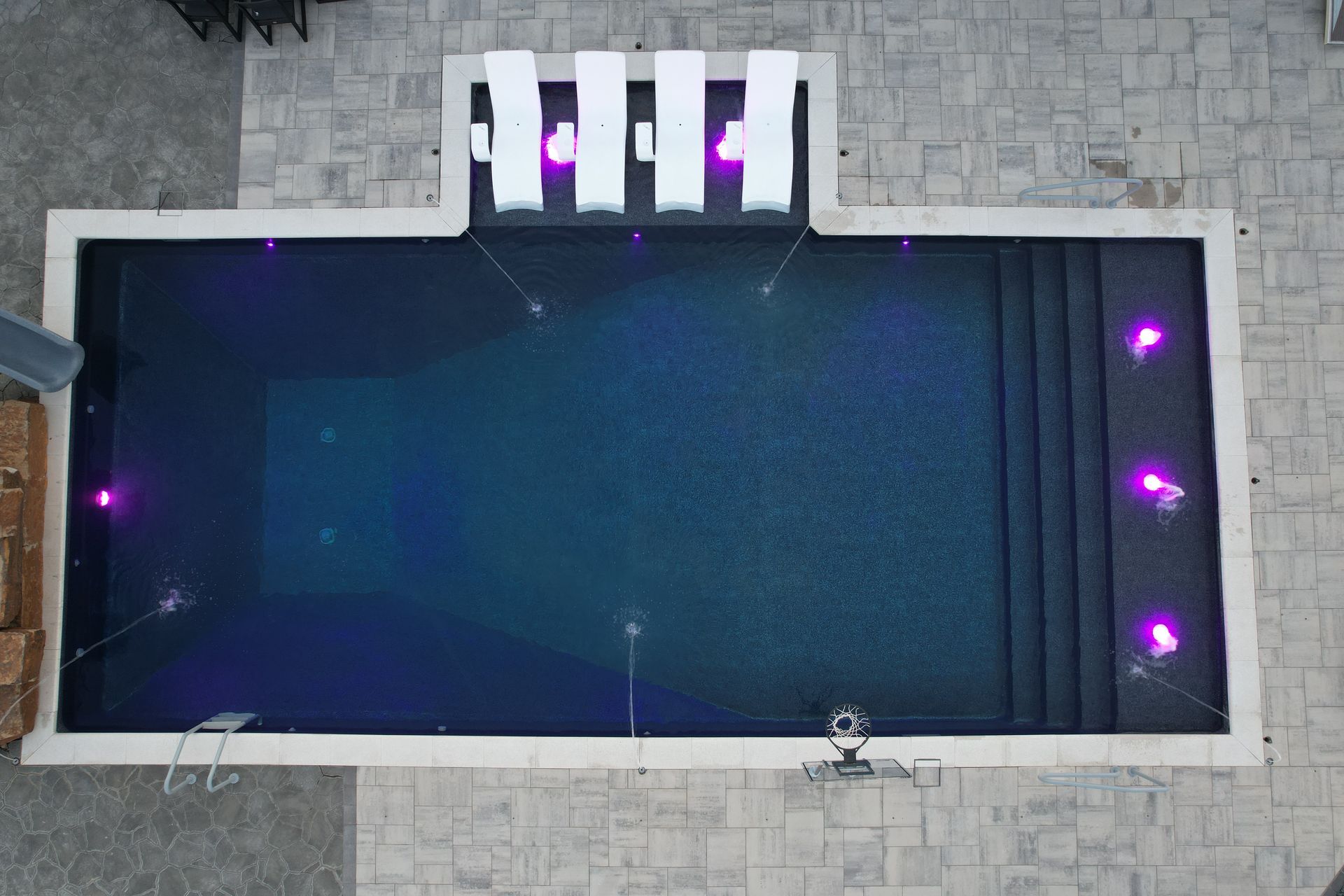 Overhead view of a rectangular pool at night, lit with purple lights and fountains; surrounding stone patio.