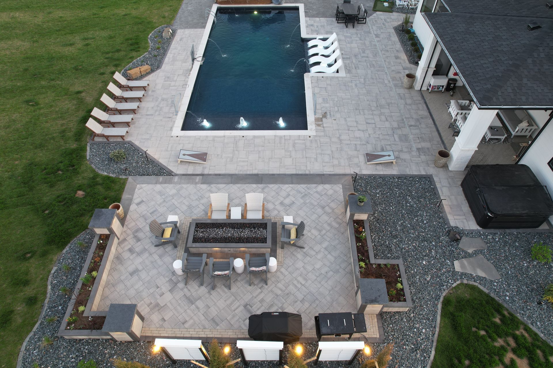 Aerial view of a backyard with a pool, patio, and outdoor kitchen. Grey pavers, dark pool water, and green lawn.