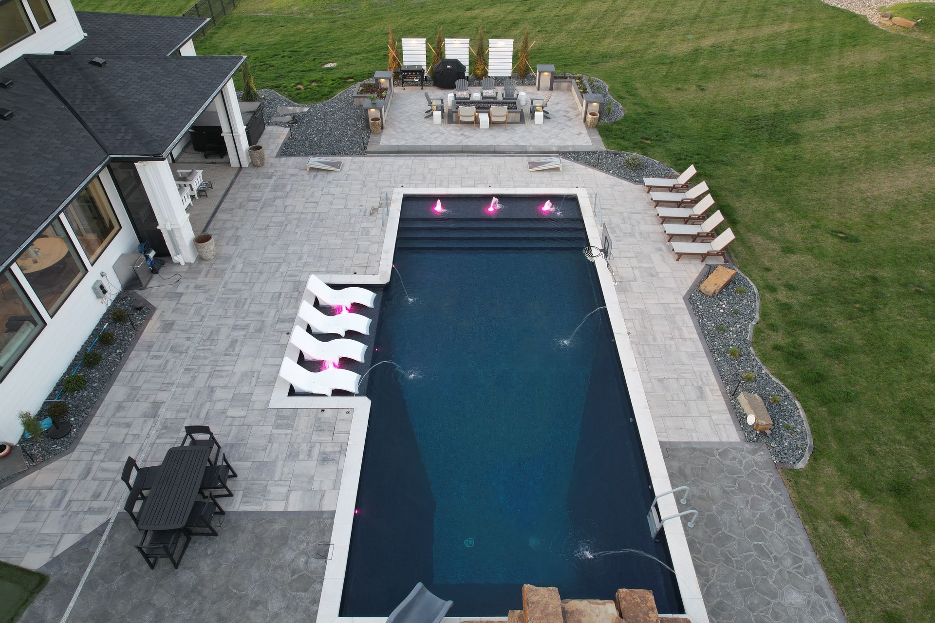 Overhead view of a rectangular pool with fountains, patio, seating area, and a house in the background.