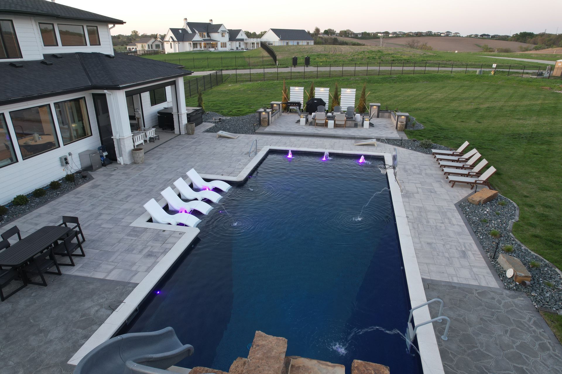 Backyard pool with white lounge chairs, slide, and patio furniture; overlooking a field.