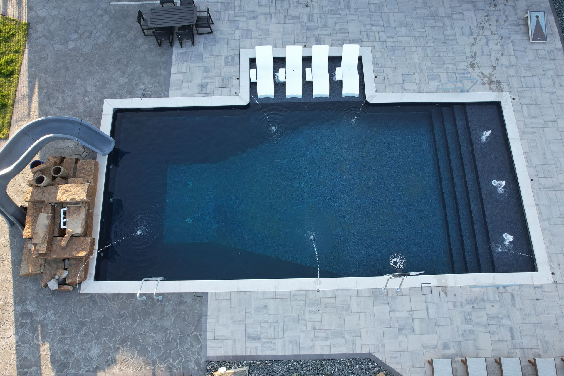Aerial view of a dark blue rectangular swimming pool with a slide and a staircase, surrounded by light gray stone.