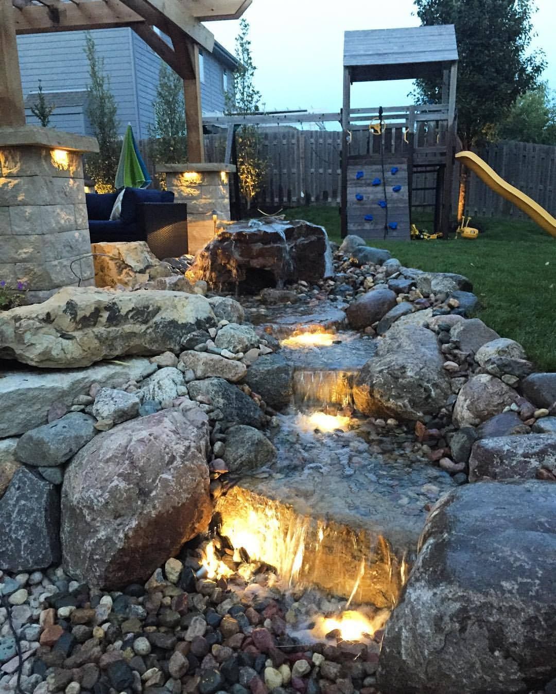 Waterfalls cascade through a rocky landscape, illuminated by soft lighting. A playset is in the background.