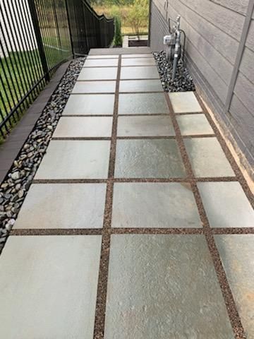 A concrete walkway with square pavers and gravel borders next to a building and fence.