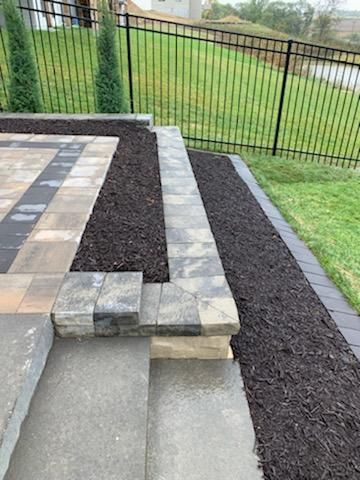 Patio with stone steps, pavers, and mulch beds alongside a grassy lawn and black fence.