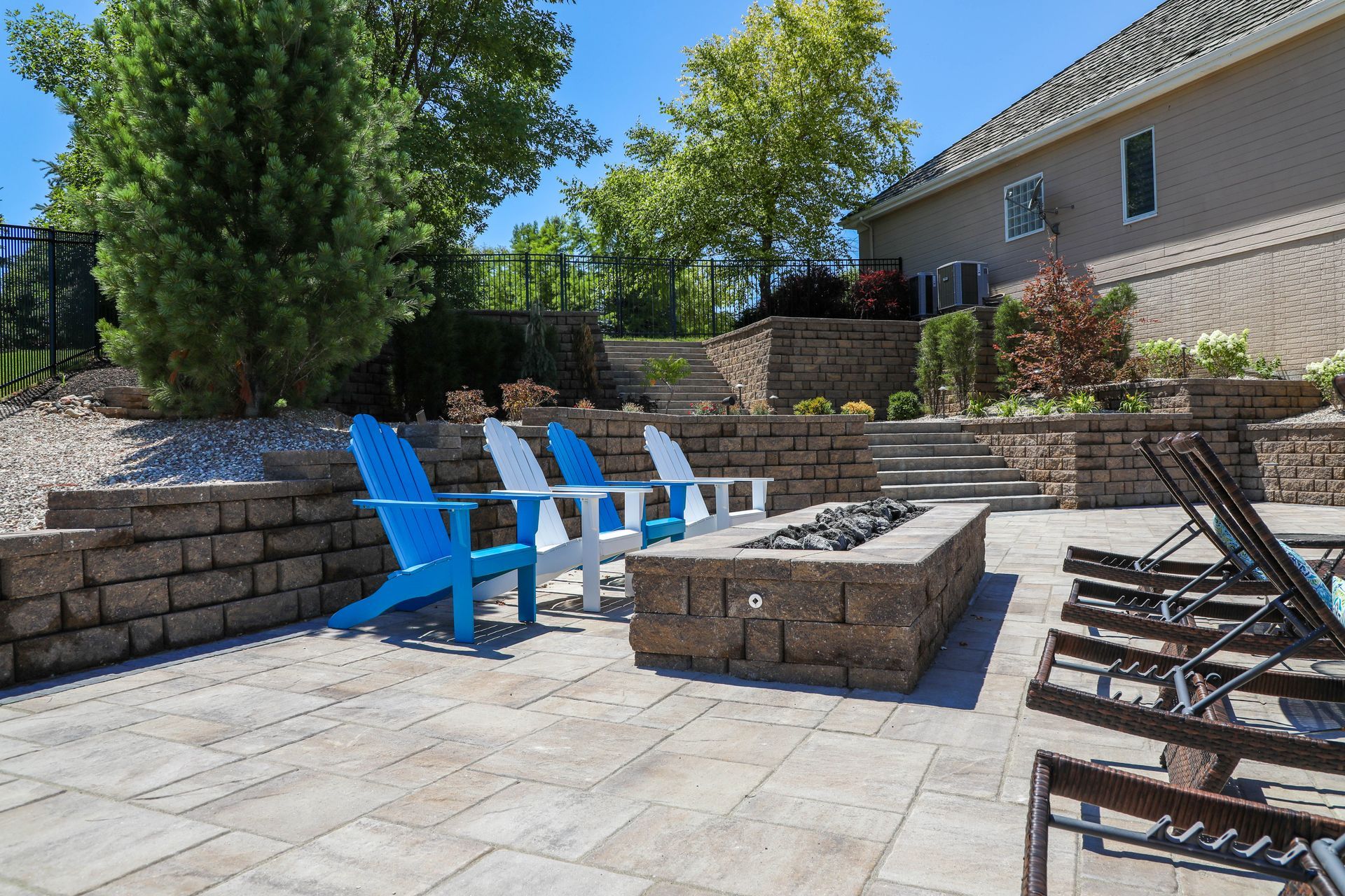 Patio with blue and white chairs, fire pit, stone walls, and a large house under a blue sky.