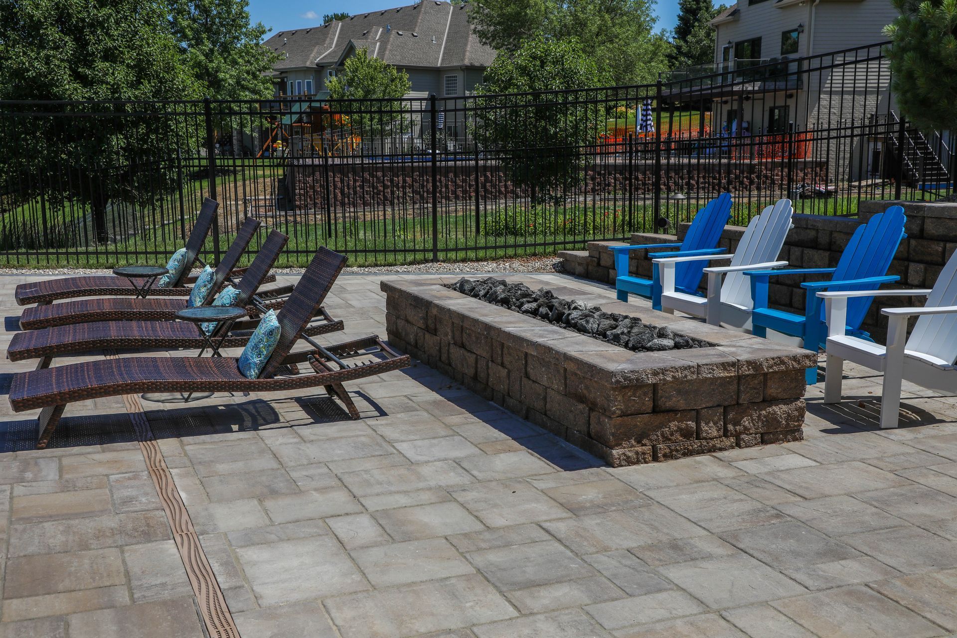 Patio with lounge chairs, blue/white Adirondack chairs around a fire pit. Black fence, buildings in background.