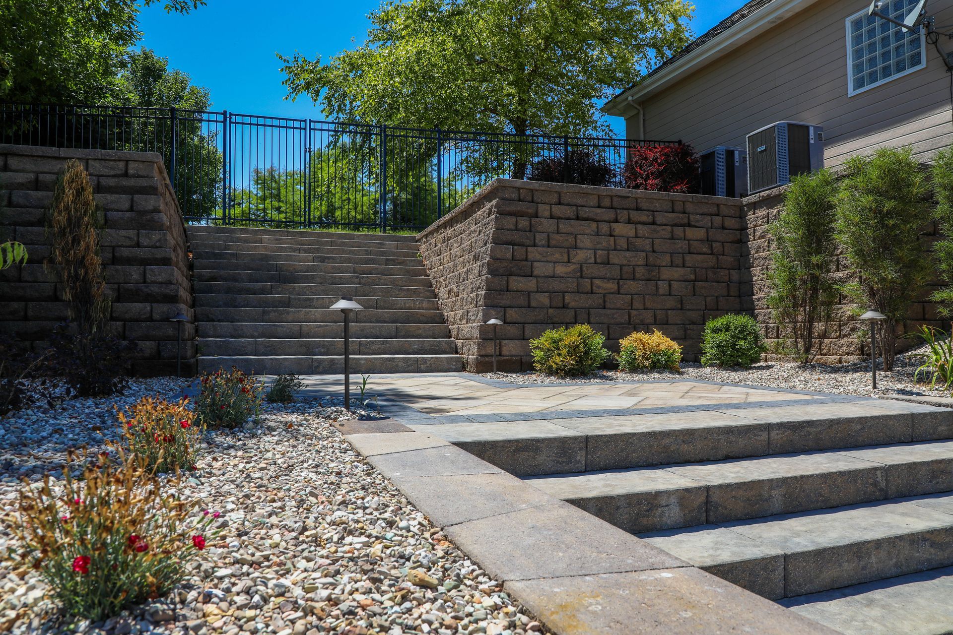 Stone steps and retaining walls in a landscaped backyard, with a black fence, gravel, and plants.