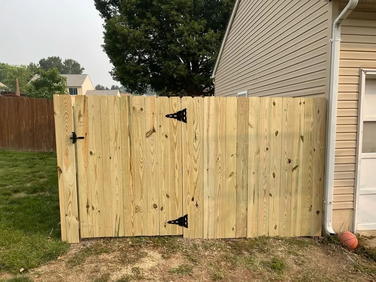 Wooden gate in backyard, attached to a house. Green grass and cloudy sky in background.