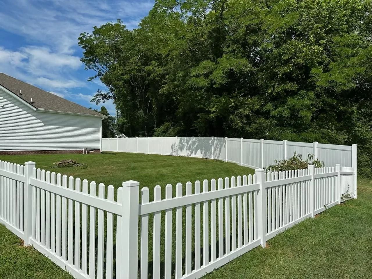 White picket fence and vinyl fence enclose a green lawn near a white house and trees under a blue sky.