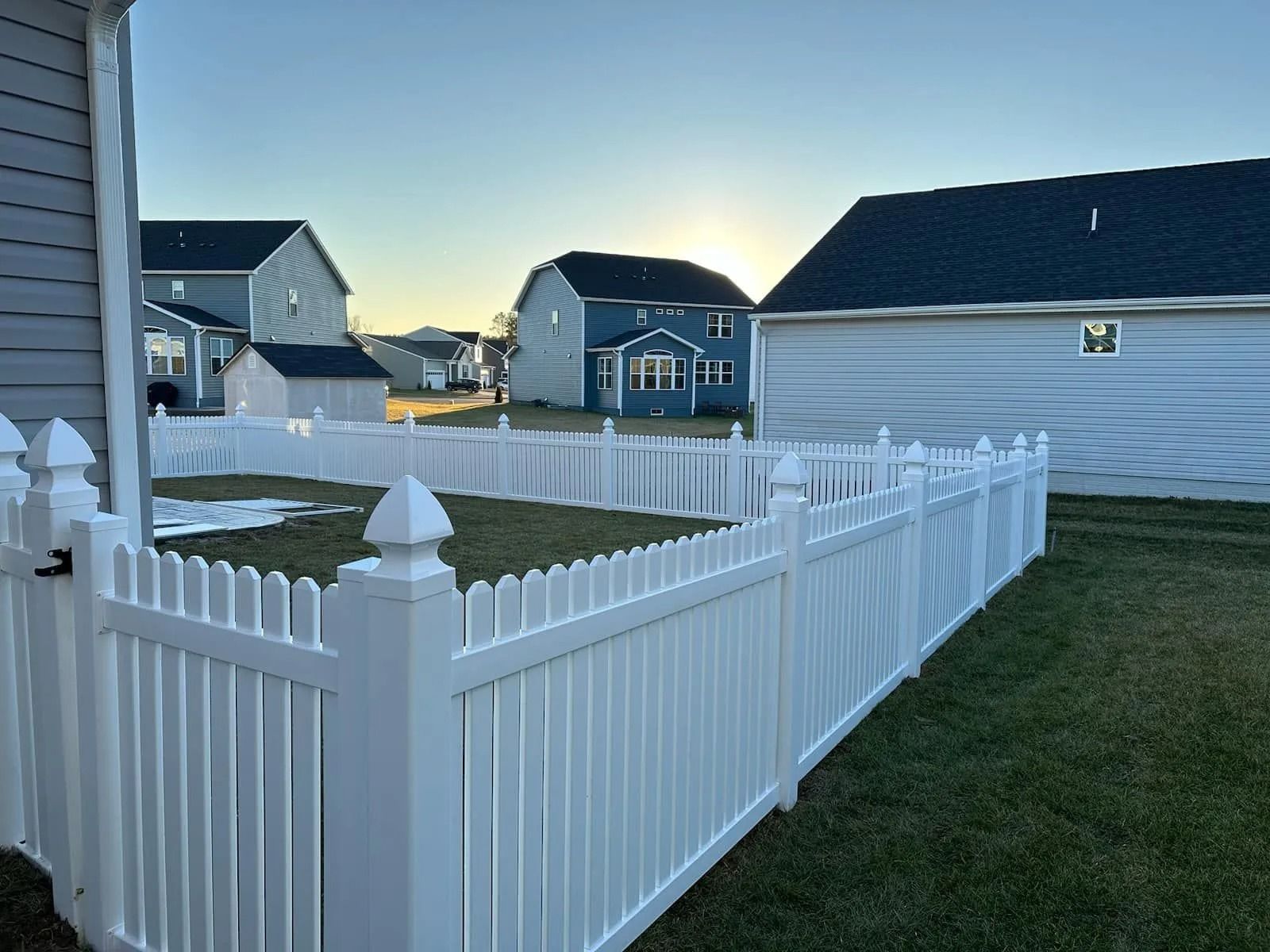 White picket fence surrounds a backyard with houses in the background under a blue sky.