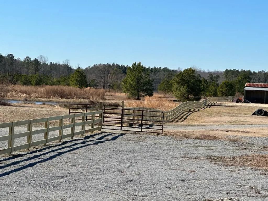 A gravel drive leads to a wooden fence and gate in a rural landscape, with trees and a small barn.