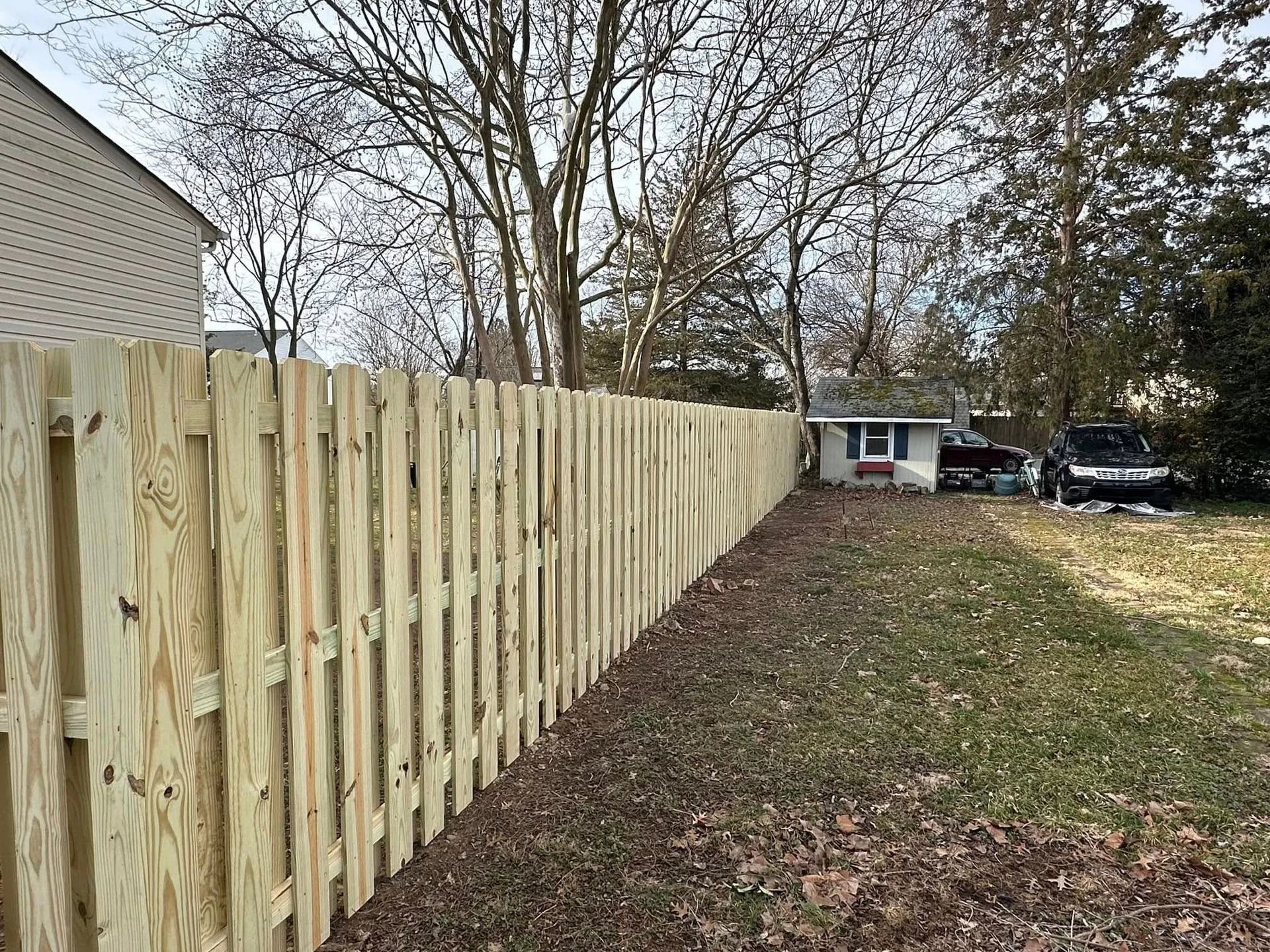 Wooden fence along a grassy yard, trees in the background, small house visible.