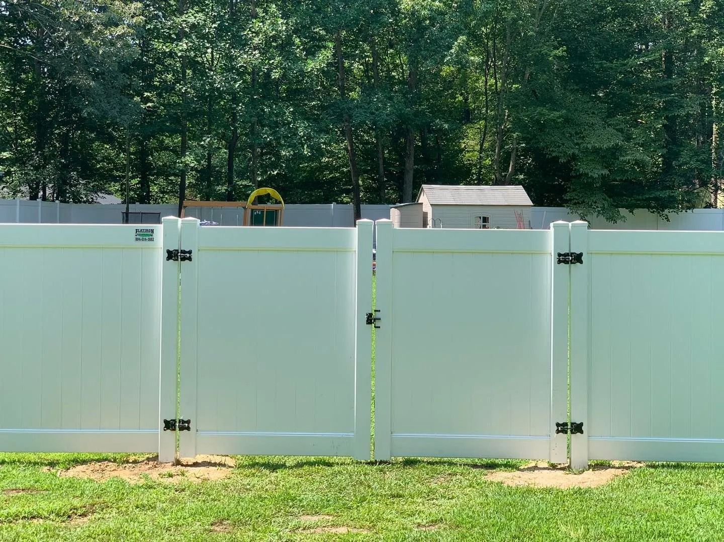 White vinyl fence with gate in a grassy yard, trees in background.