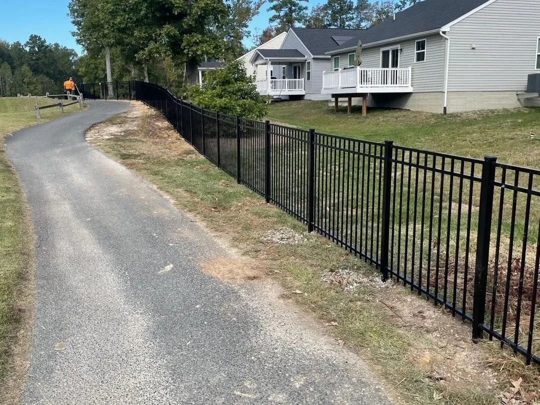 Black metal fence alongside a paved path curving through grassy land, houses in the background.