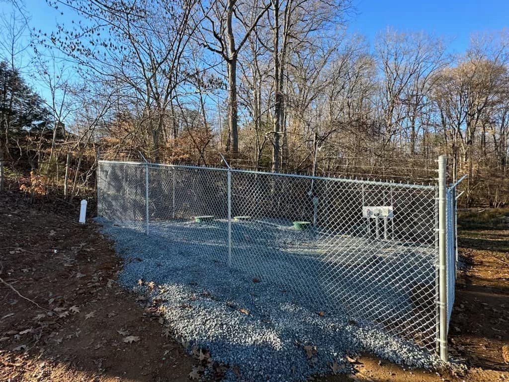 Chain-link fence enclosure with gravel base, surrounded by trees. Sunny day.
