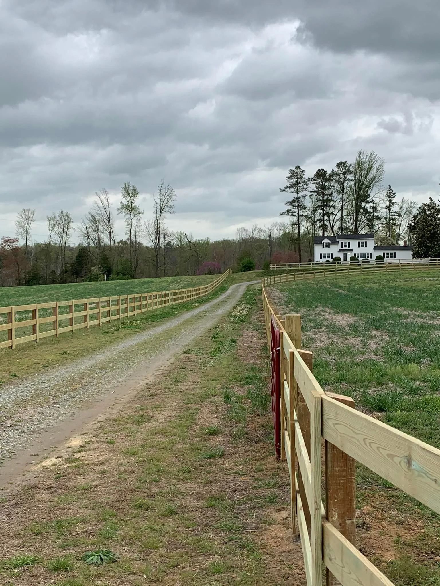 Gravel driveway leading to a white house, flanked by wooden fences. Cloudy sky, grassy field, trees in the background.
