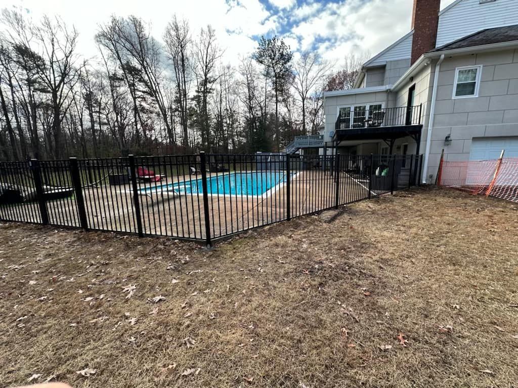 Black fenced-in swimming pool next to a house with a deck; brown grass and trees in the background under a cloudy sky.