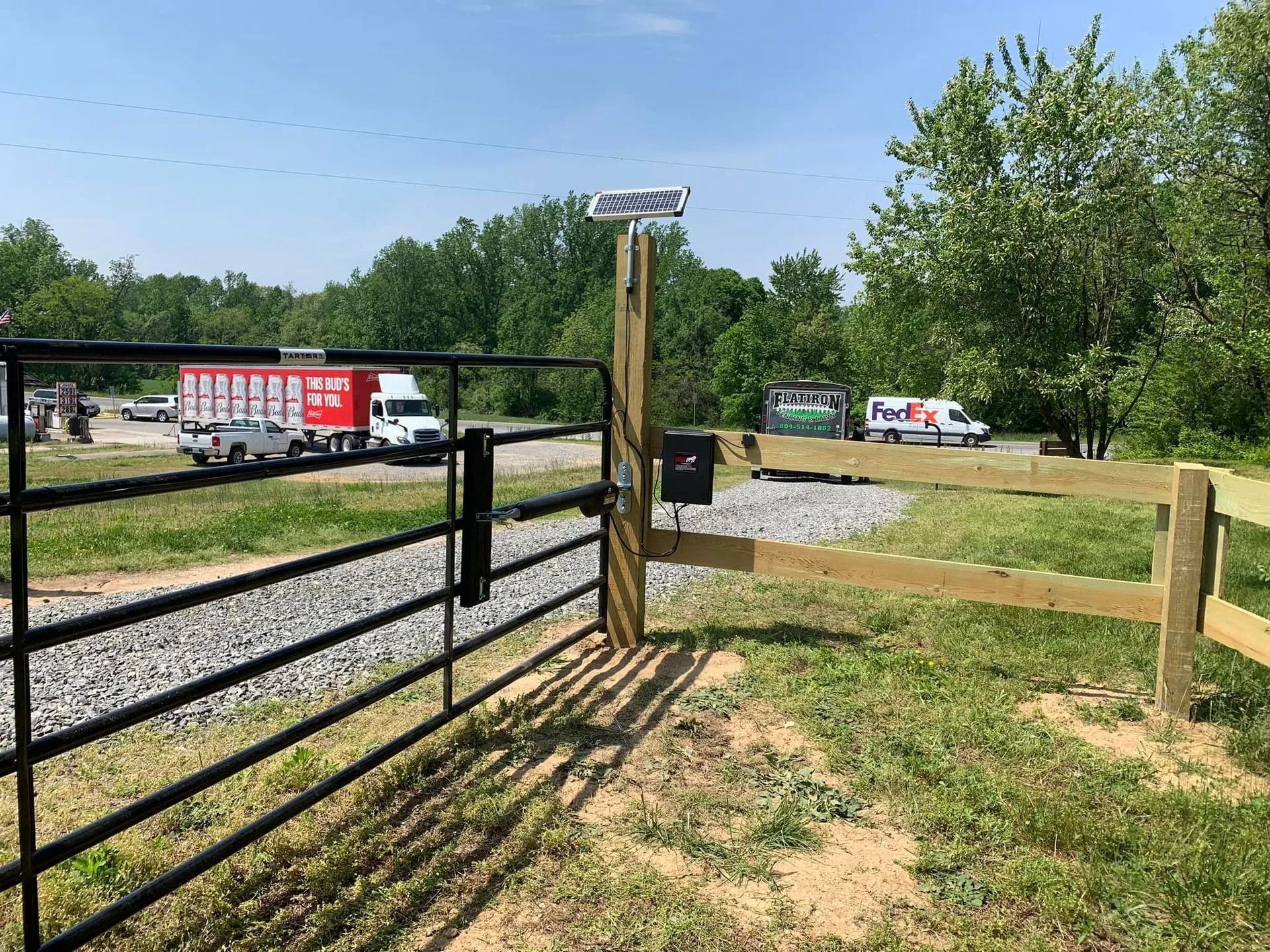 Black gate, wooden post with solar panel, gravel driveway, vehicles in background, and green grass.