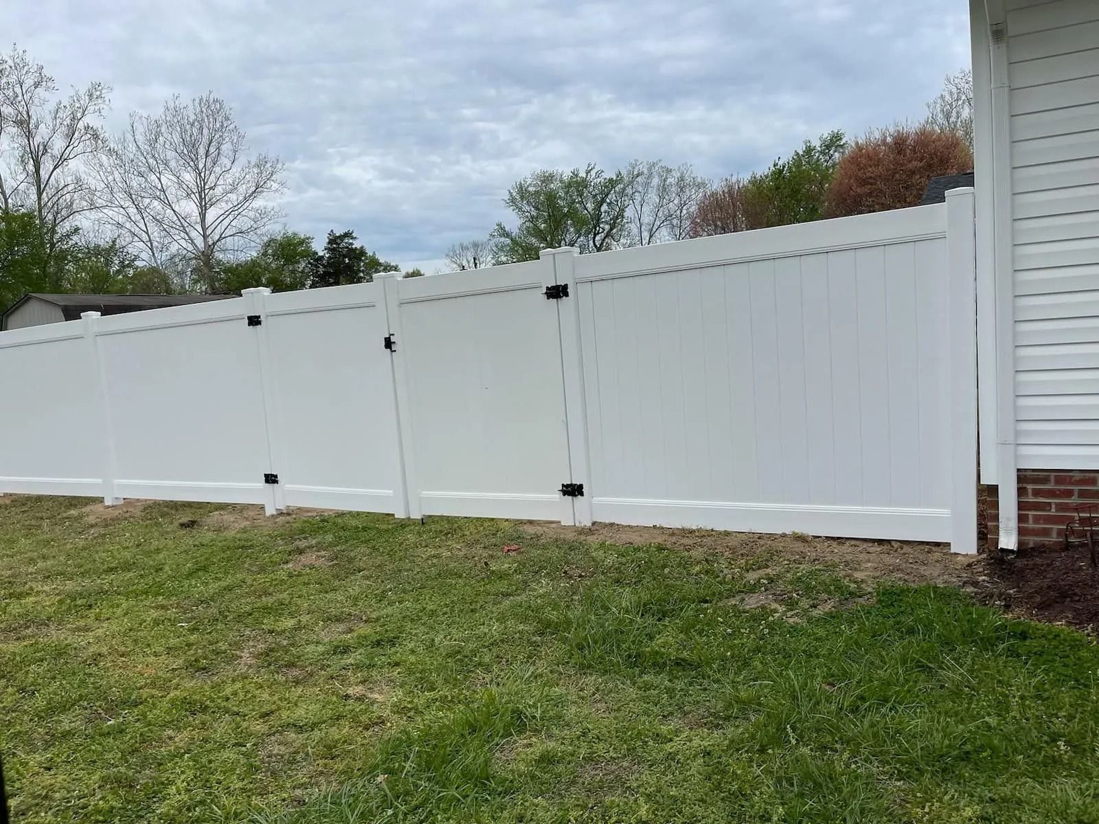 White vinyl privacy fence in a backyard, with two gates and a house on the right.