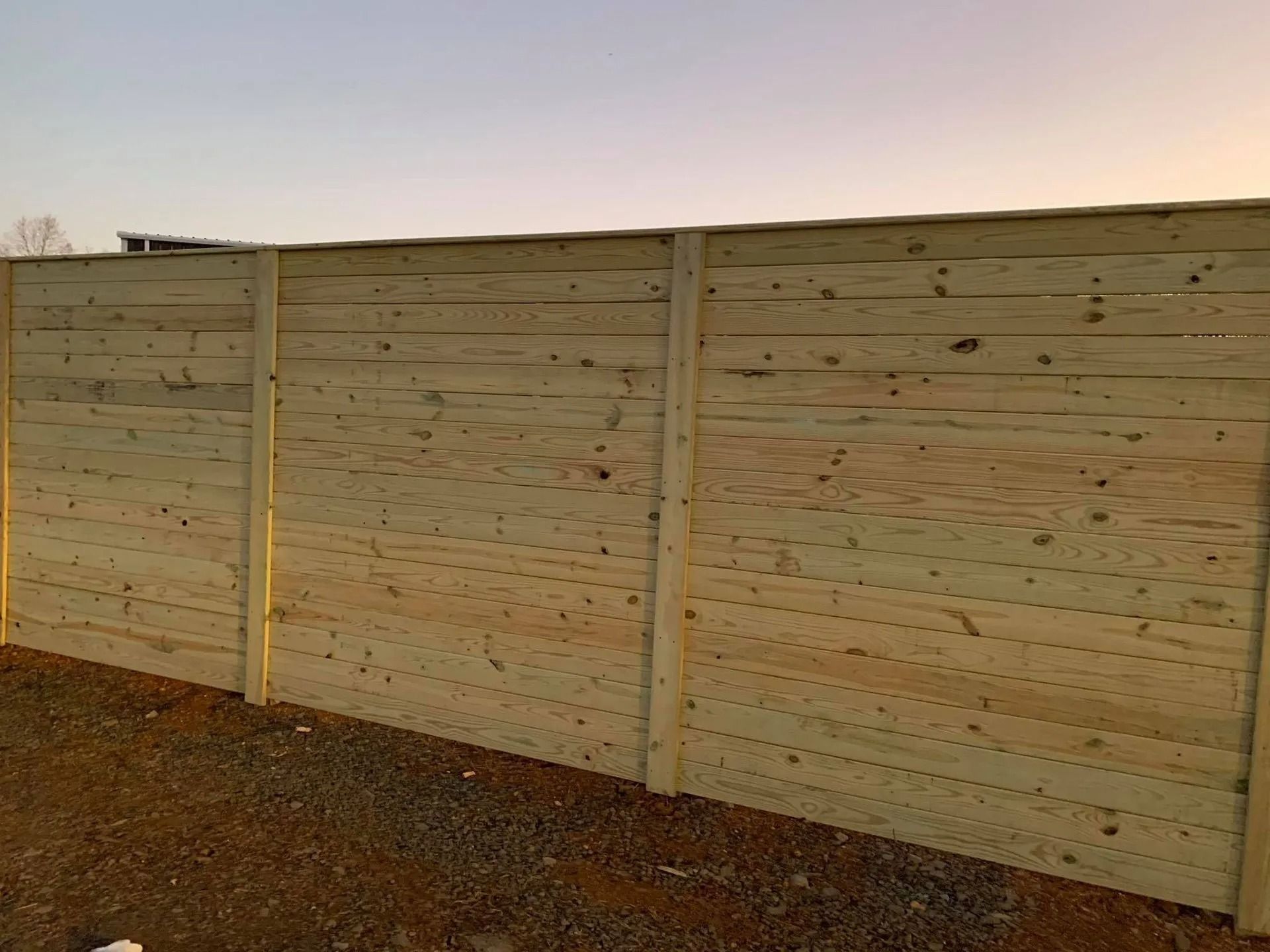 Wooden privacy fence with horizontal planks, posts set on gravel ground, under a dusky sky.