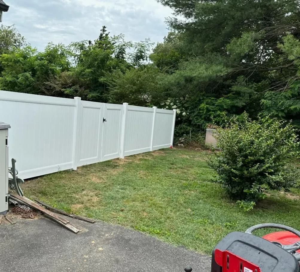 White fence in a yard with green grass, bushes, and trees. A red lawnmower is on the right.