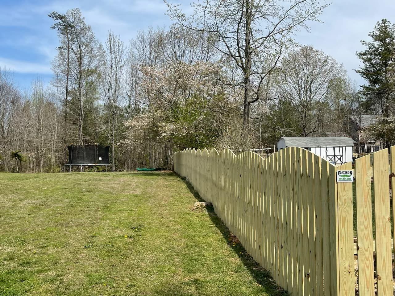 Wooden fence in a grassy yard, with trees and a shed in the background under a blue sky.