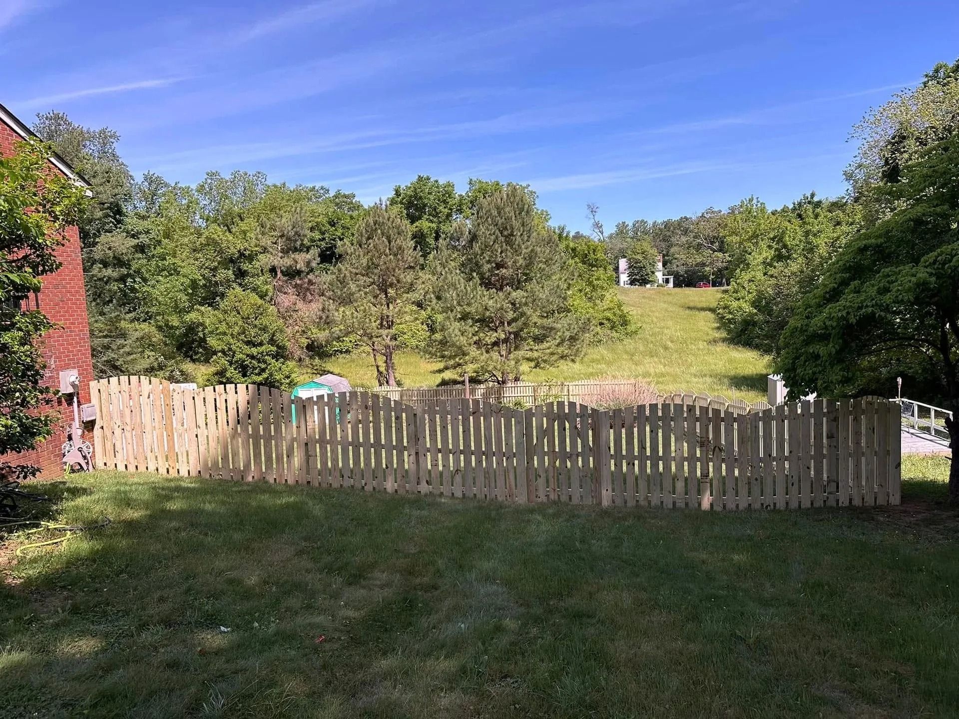 Wooden fence in a grassy yard, with trees and a hillside in the background under a blue sky.