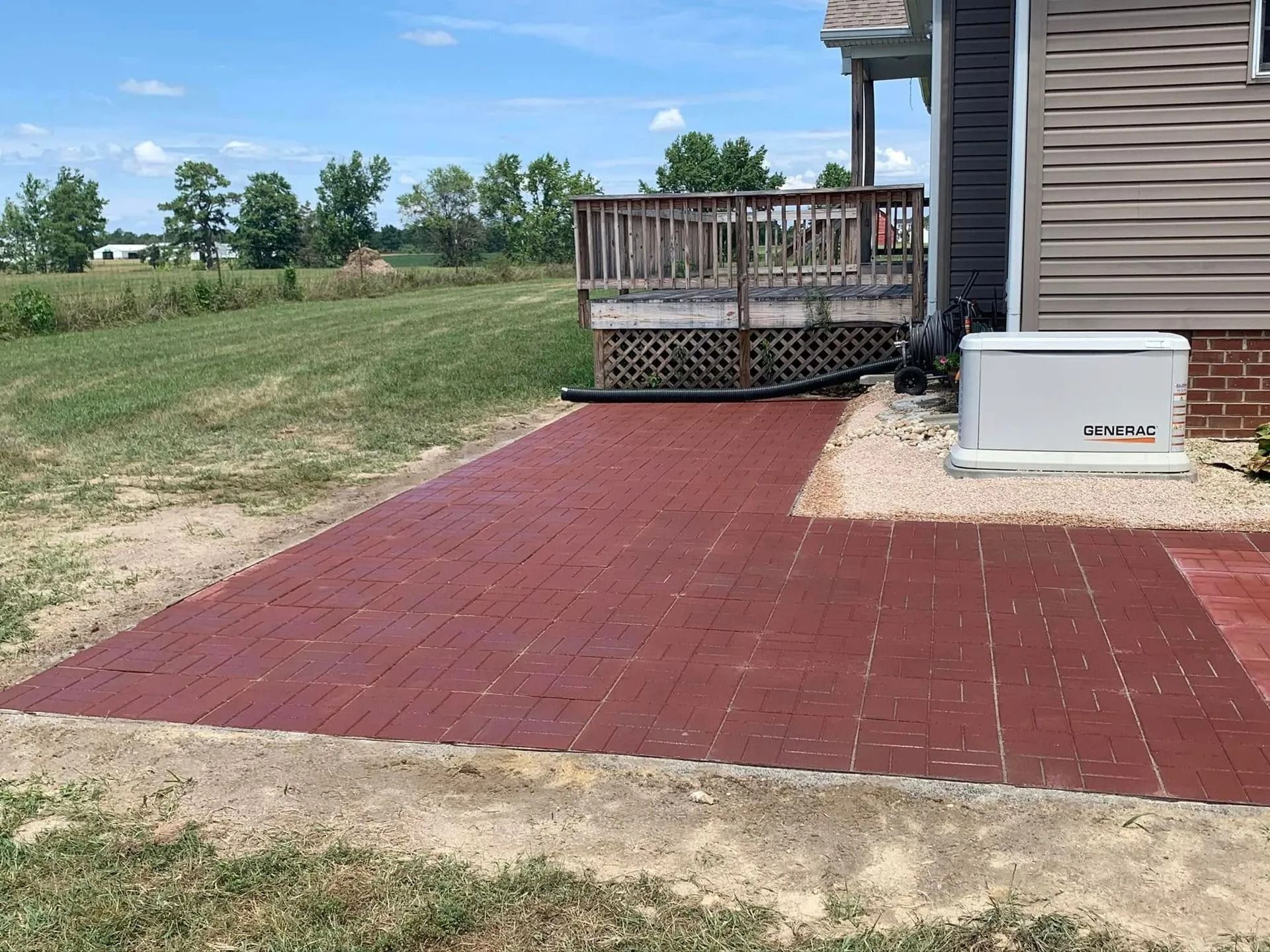 Red brick patio next to a wooden deck, a generator, and a house with brown siding, outdoors.