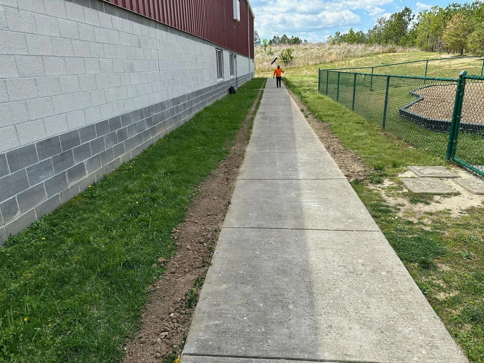 A concrete path beside a gray brick building, grass on either side, and a person walking in the distance.