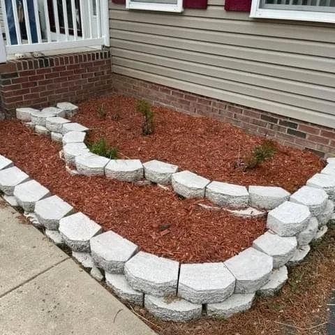 A low garden bed with gray stone blocks filled with brown mulch and small plants, next to a beige house.