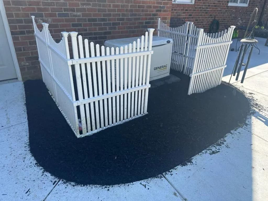 White fenced enclosure around a generator on black rubber mulch next to a brick building.