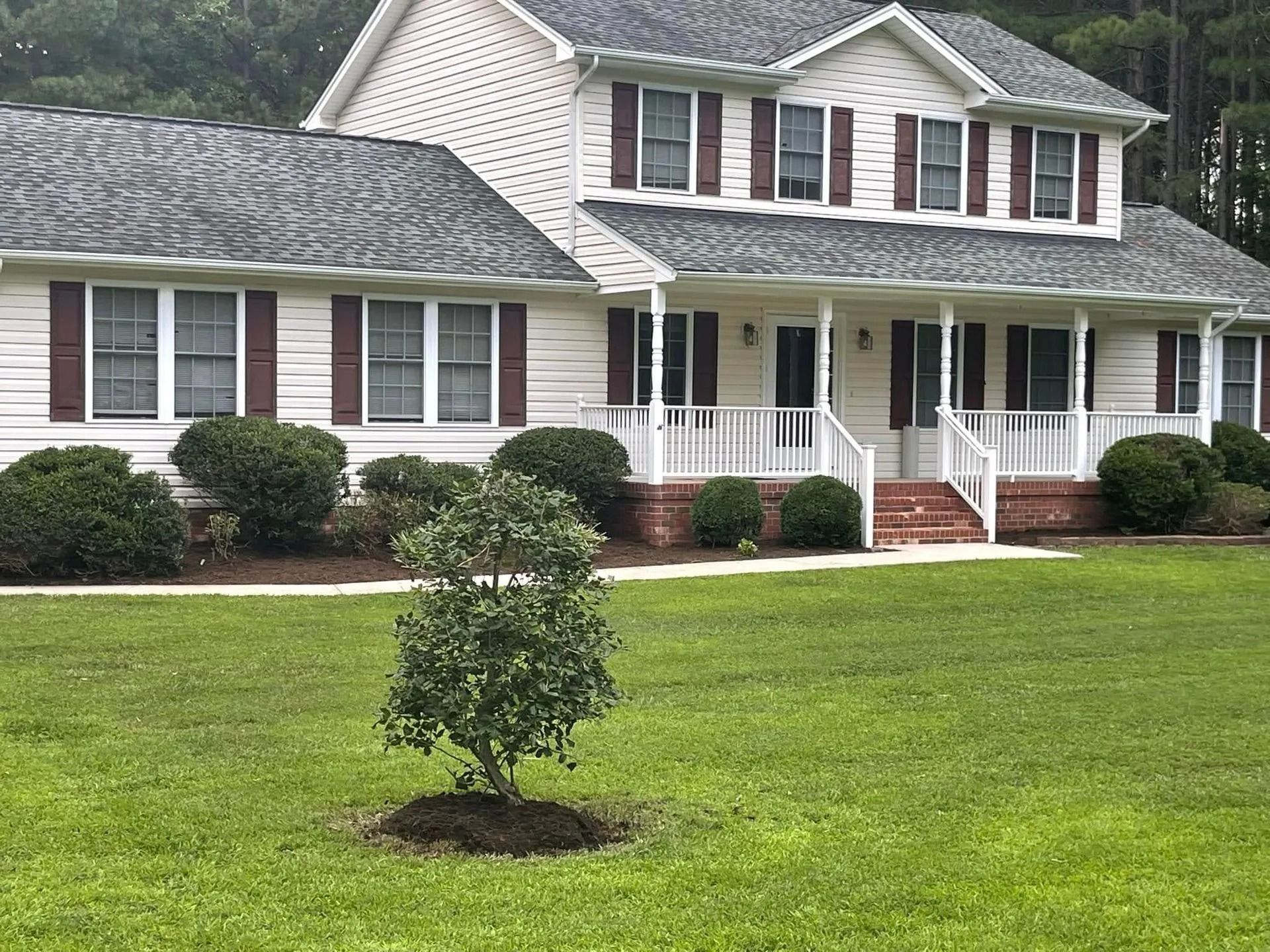 Two-story white house with maroon shutters and a front porch. Green lawn with a small tree in the foreground.