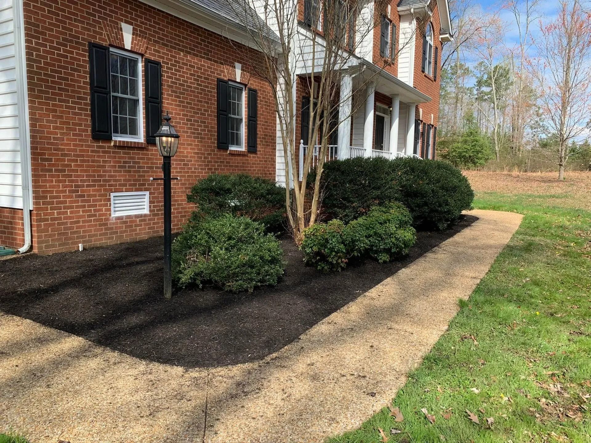 A brick house with black shutters and a freshly mulched garden bed. A dark lamppost stands near the bushes.