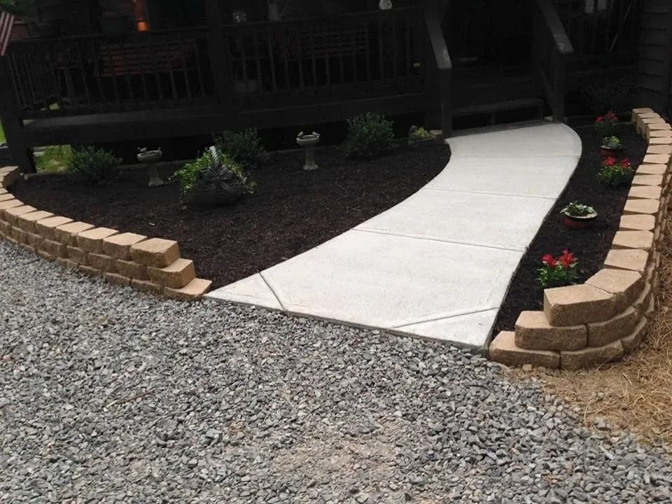 Concrete walkway leading to a dark wooden porch, bordered by retaining walls and landscaping with mulch and flowers.