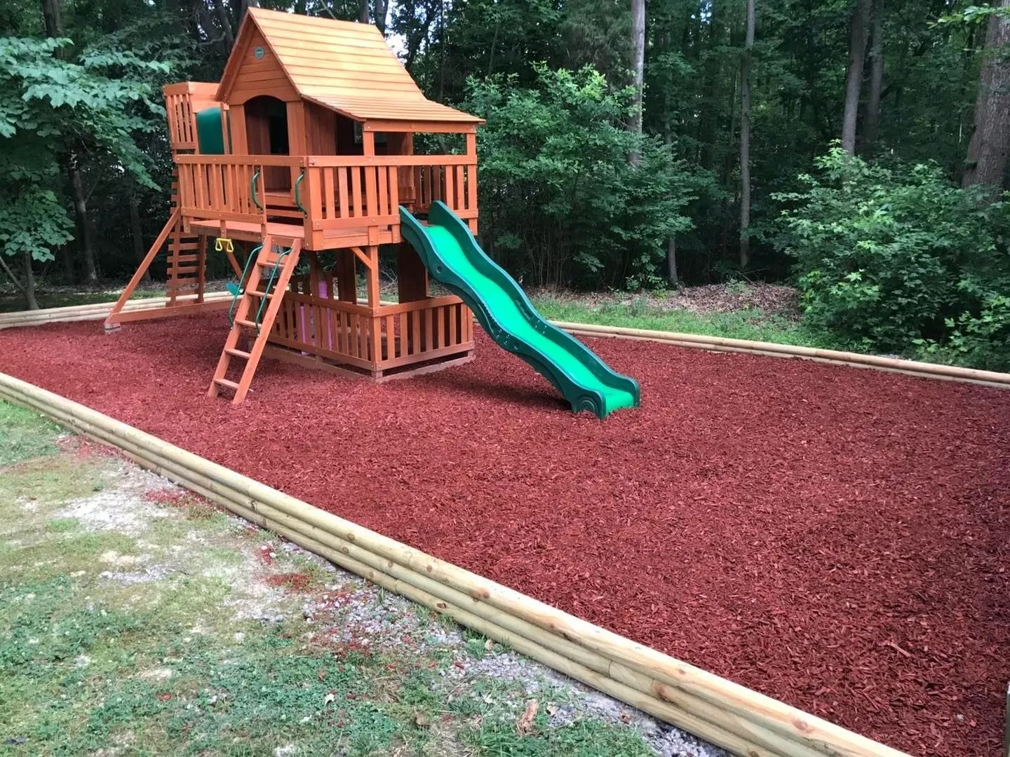 Wooden playset with a green slide on red mulch, bordered by logs, in a grassy backyard.
