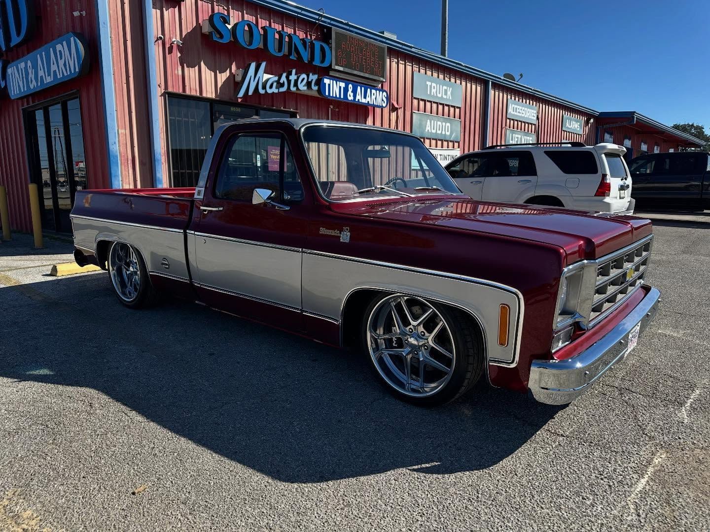 Maroon-and-cream classic pickup truck parked outside a rustic shop on a sunny day