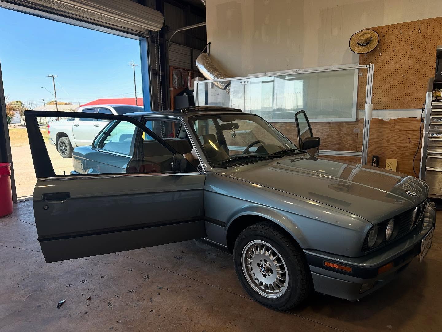 Gray vintage car with open driver’s door inside a garage, with another car visible outside.