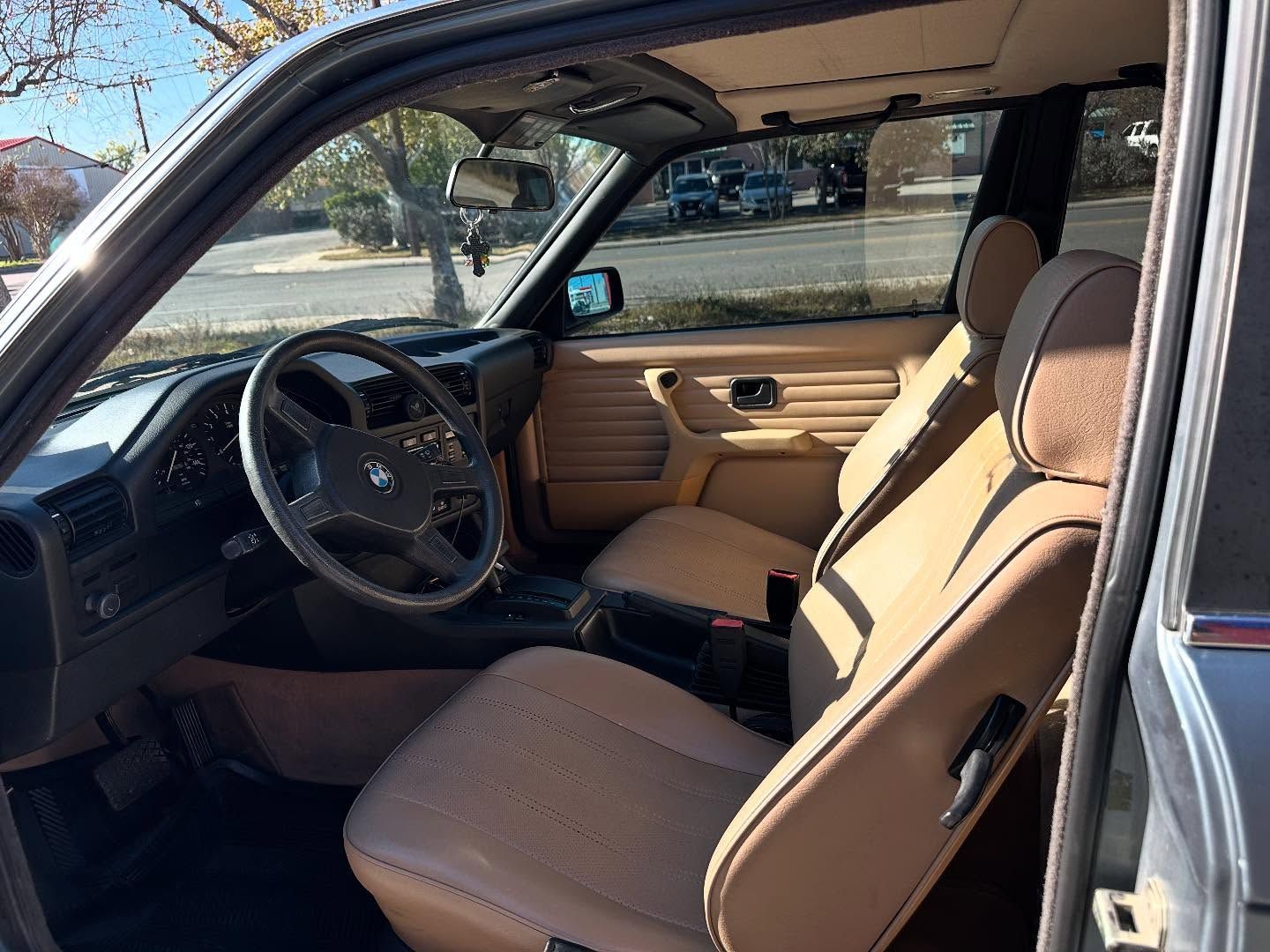 Car interior with tan leather seats and black steering wheel, viewed through the open driver’s door.