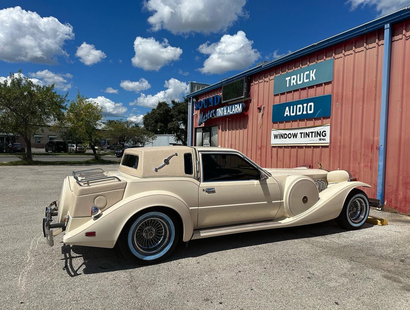 Cream vintage convertible parked beside an audio truck sign under a bright blue sky.