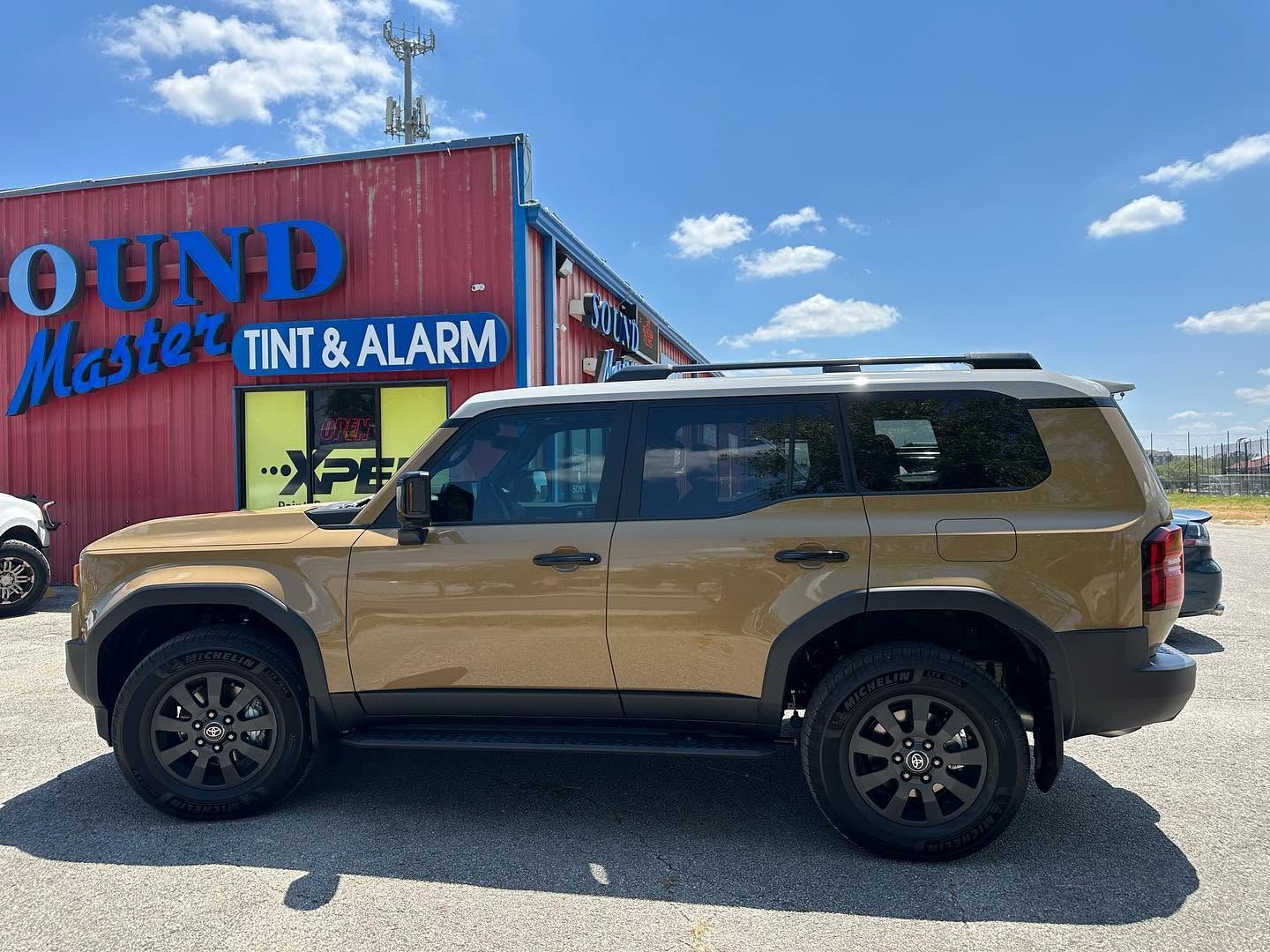 Tan SUV parked outside a building with a blue sign reading “TINT & ALARM” under a sunny sky
