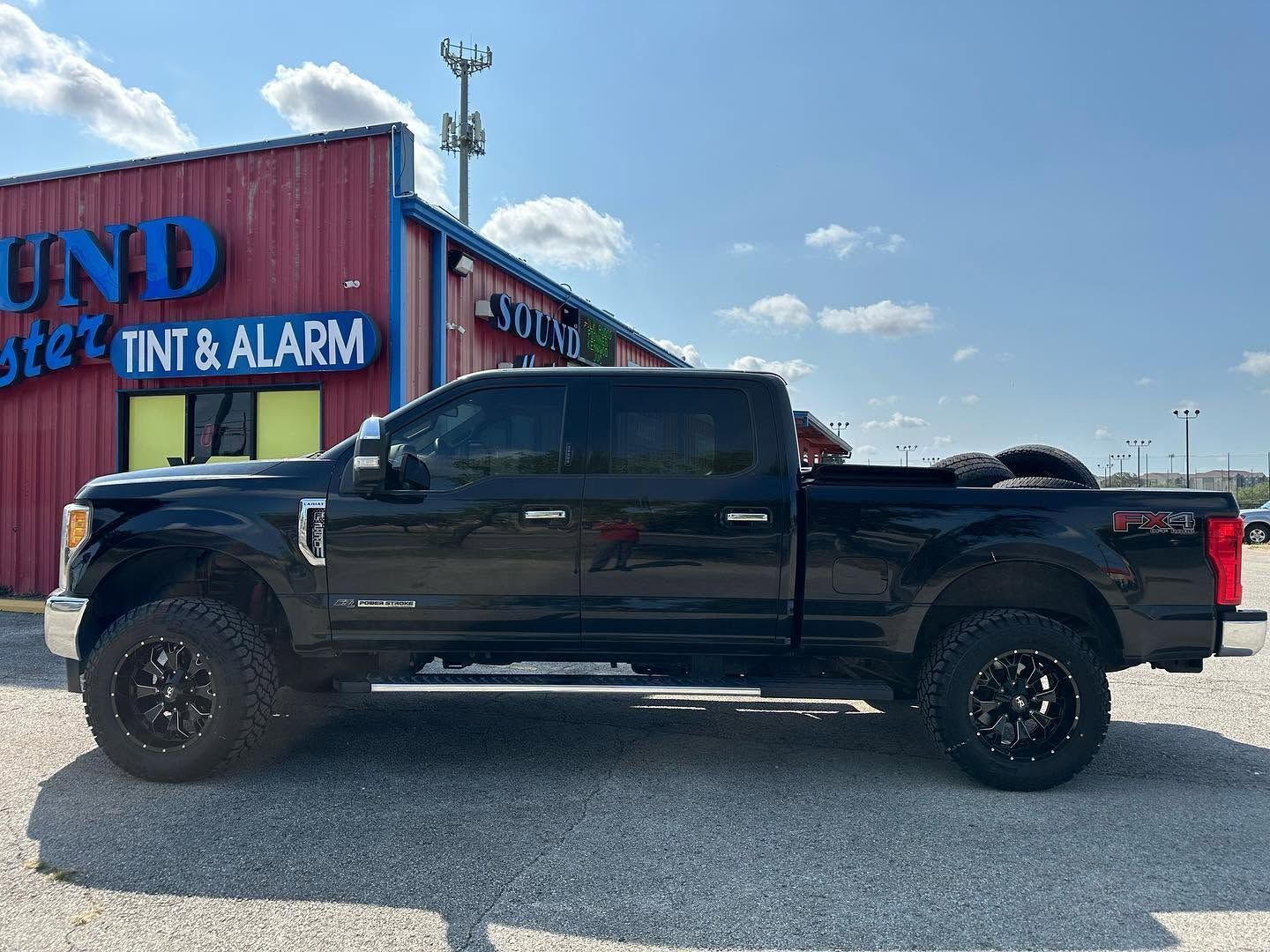 Black pickup truck parked beside a red building on a gravel lot under a blue sky.