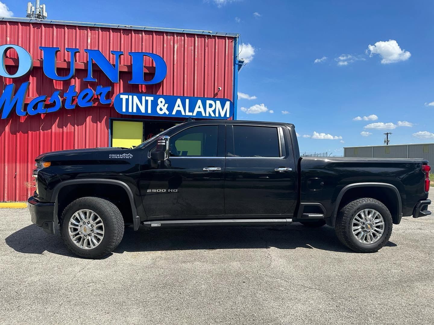 Black pickup truck parked beside a red building with “Tint & Alarm” signage under a blue sky
