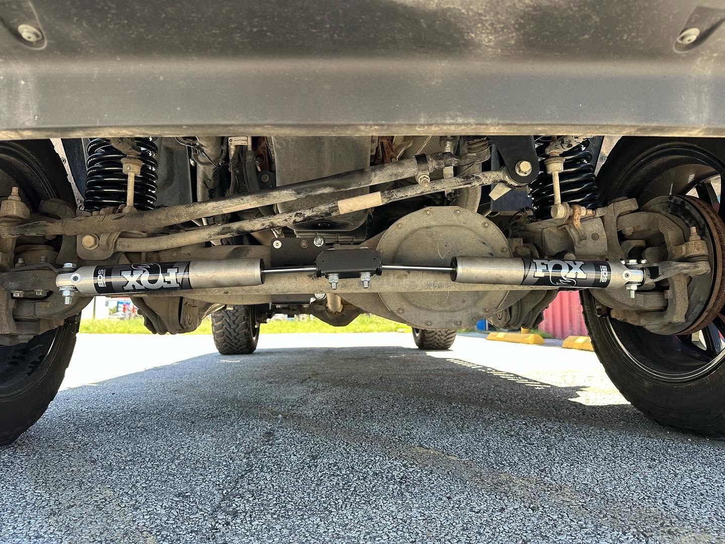 Underside of a vehicle showing the front axle, suspension, and steering components on gravel.