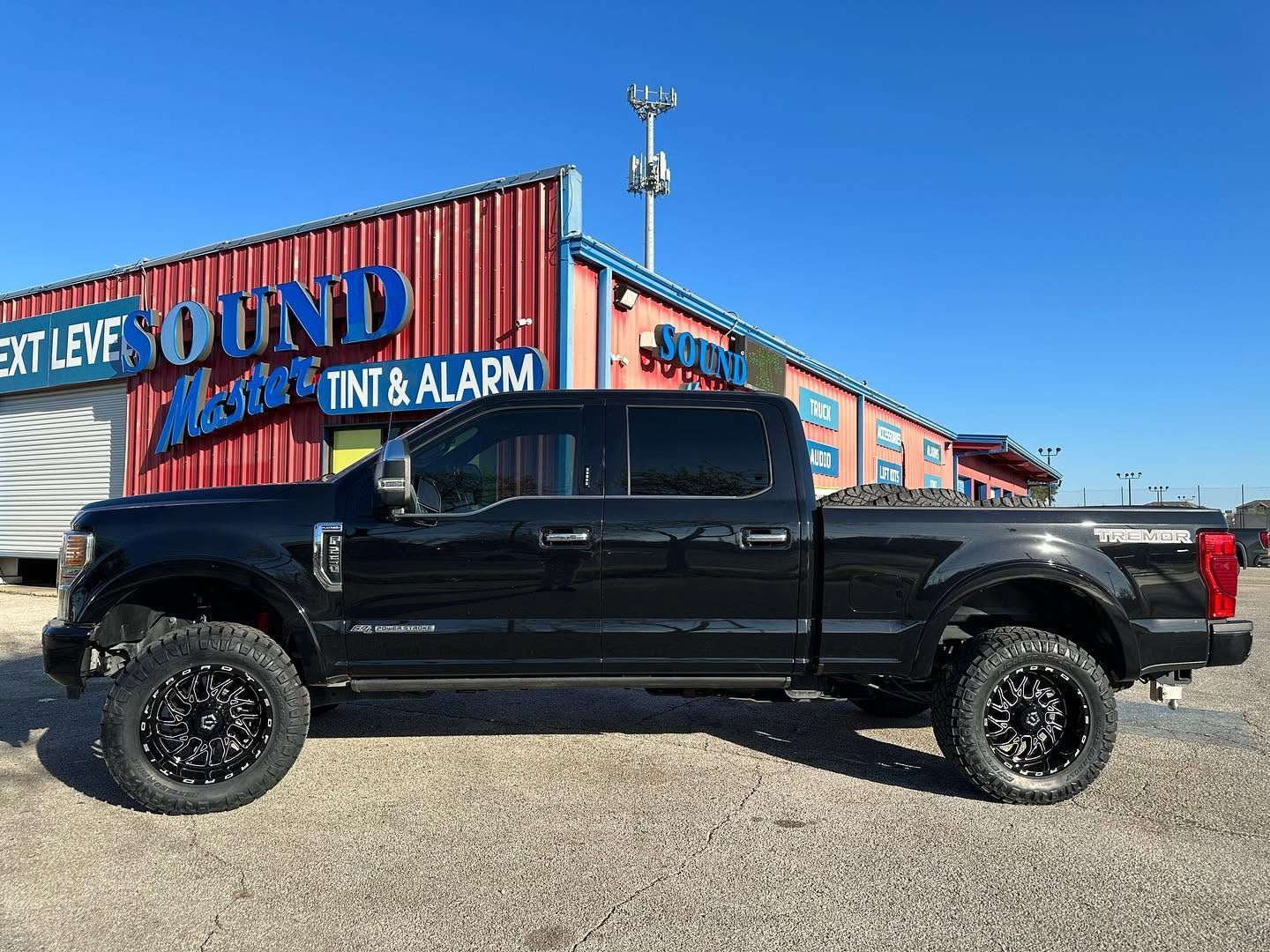 Black lifted pickup truck parked outside a sound, tint, and alarm shop under a blue sky.